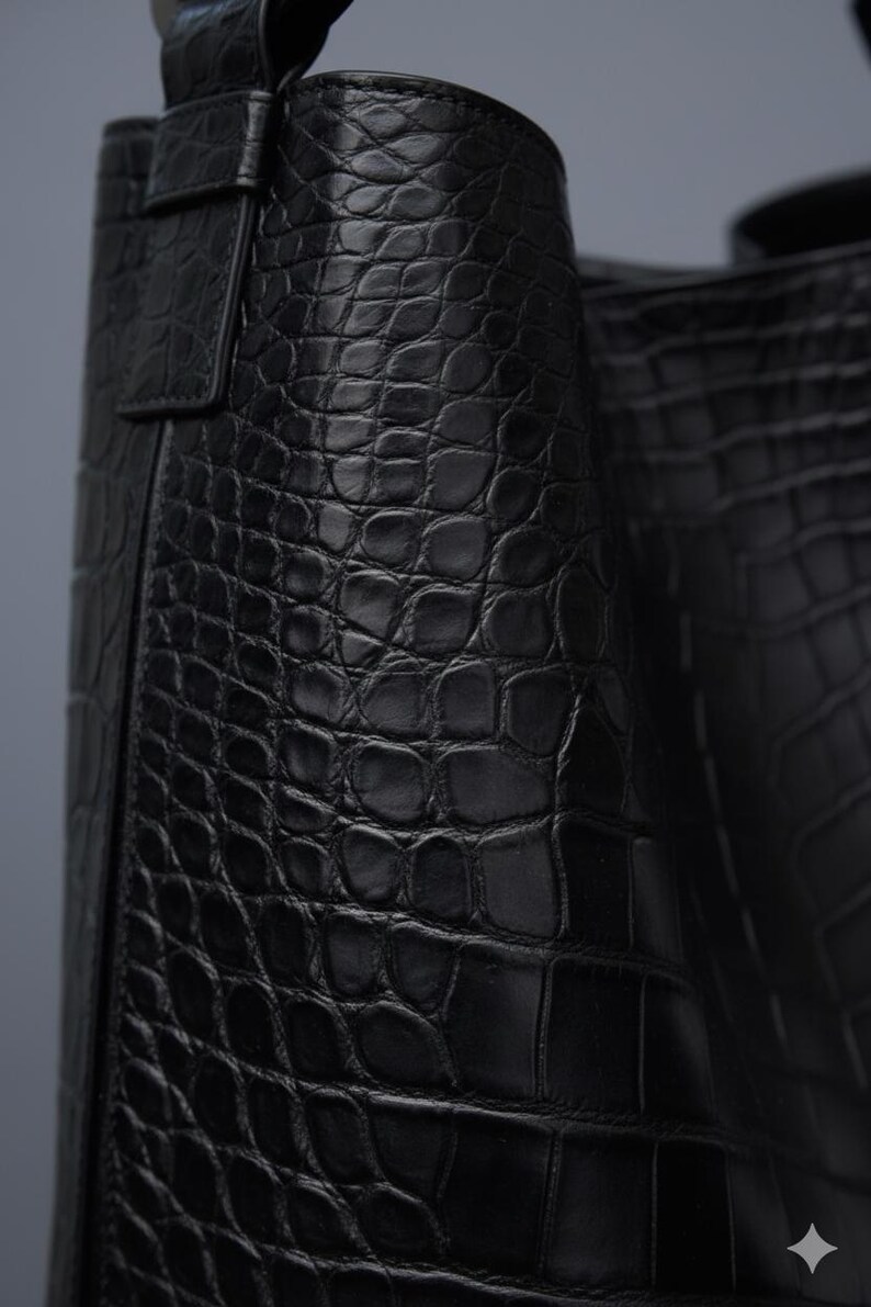 K&ouml;nnte beinhalten: Nahaufnahme einer schwarzen Lederhandtasche mit Krokodillederstruktur. Die Tasche hat ein strukturiertes Design mit einem Griff und einem Klappenverschluss. Das Leder scheint hochwertig zu sein und die Tasche ist wahrscheinlich ein Luxusartikel.