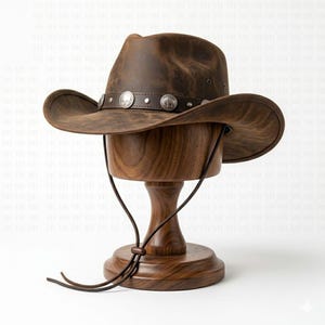 May include: A brown leather cowboy hat featuring a dark brown leather hatband adorned with silver-toned metal conchos depicting buffalo figures. The hat includes a thin brown leather chin cord with an adjustable sliding bead for secure fastening.