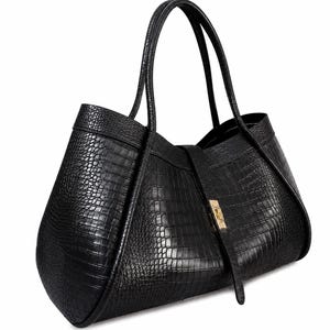 May include: A black tote bag featuring a textured crocodile skin pattern, two thin shoulder straps, and a central vertical strap closure secured with a small gold-toned metal clasp. The bag has a wide, structured base and curved side panels.