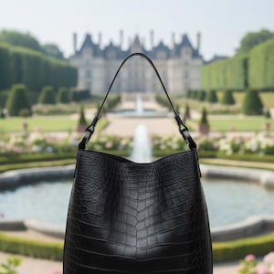 K&ouml;nnte beinhalten: Eine schwarze Tragetasche mit Krokodil-Textur. Die Tasche hat einen einzelnen Henkel und eine strukturierte Form. Der Hintergrund zeigt einen verschwommenen Garten mit einem Brunnen und einem Geb&auml;ude.