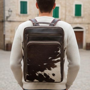 Puede incluir: Una mochila de cuero marrón con un bolsillo frontal de piel de vaca. La mochila tiene un asa superior, correas ajustables y un compartimento principal con cremallera. El bolsillo de piel de vaca es marrón y blanco.