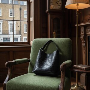 K&ouml;nnte beinhalten: Eine schwarze Ledertasche mit Krokodiltextur liegt auf einem gr&uuml;nen Sessel. Der Sessel hat Holzarmlehnen. Eine Standuhr und eine Stehlampe befinden sich im Hintergrund. Der Raum hat ein klassisches Design.