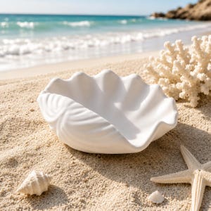 May include: A matte white ceramic bowl shaped like a large clam shell with a scalloped edge and textured exterior ridges. The bowl is positioned on a sandy surface alongside a piece of white coral, a small spiral seashell, and a five-pointed starfish.