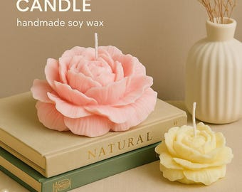 Könnte beinhalten: Eine rosa Pfingstrosen-Blütenkerze aus handgemachtem Sojawachs liegt auf Büchern. Daneben befindet sich eine kleinere gelbe Rosenkerze. Der Text "PEONY FLOWER CANDLE" und "handmade soy wax" sind sichtbar.