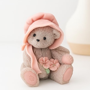 May include: A detailed figurine of a teddy bear wearing a pink bonnet and scarf, holding pink flowers. The bear has a light brown fur texture, with pink accents on its paws and ears. The background is a soft white.