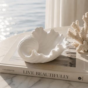 Może przedstawiać: Biała ceramiczna miska w kształcie muszli ze spiralnym wzorem, spoczywająca na książce z napisem "LIVE BEAUTIFULLY". Obok miski znajduje się biała dekoracja z koralowców. Tło ukazuje fragment oceanu.