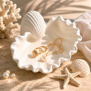 May include: A white ceramic shell-shaped trinket dish holding two gold rings and a gold chain necklace with a pearl pendant. The dish features a scalloped edge and a ribbed exterior, resting alongside a starfish, a small seashell, and loose pearls.