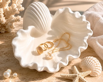May include: A white ceramic shell-shaped trinket dish holding two gold rings and a gold chain necklace with a pearl pendant. The dish features a scalloped edge and a ribbed exterior, resting alongside a starfish, a small seashell, and loose pearls.