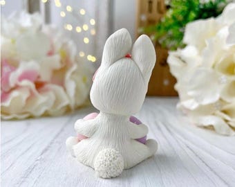 May include: A white ceramic bunny figurine, viewed from the back, holding two pastel Easter eggs. The bunny has a small pink bow and a textured, fluffy tail. The figurine is on a white wooden surface, with flowers in the background.