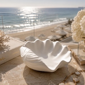 May include: A matte white ceramic bowl shaped like a large clam shell with scalloped edges and a textured exterior. The bowl is positioned centrally, featuring a smooth interior basin and a detailed, ridged outer surface that mimics natural shell anatomy.