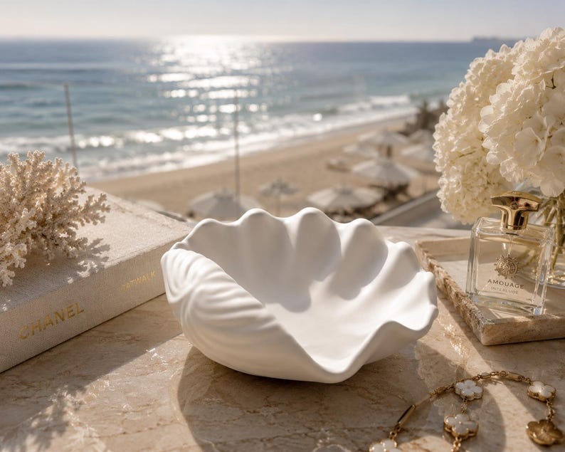 May include: A matte white ceramic bowl shaped like a large clam shell with scalloped edges and a textured exterior. The bowl is positioned centrally, featuring a smooth interior basin and a detailed, ridged outer surface that mimics natural shell anatomy.
