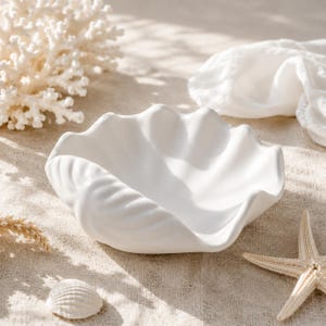 May include: A matte white ceramic bowl shaped like a large clam shell with deep, undulating ridges and a smooth, curved rim. The bowl features a wide, open basin and a textured exterior that mimics the natural form of a seashell, finished in a clean, solid white color.