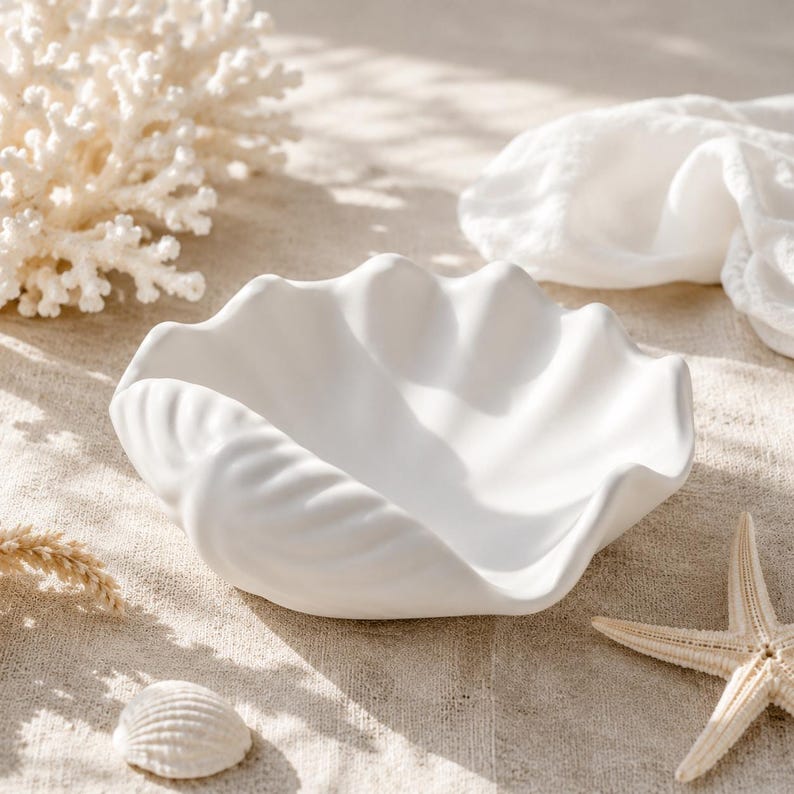 May include: A matte white ceramic bowl shaped like a large clam shell with deep, undulating ridges and a smooth, curved rim. The bowl features a wide, open basin and a textured exterior that mimics the natural form of a seashell, finished in a clean, solid white color.