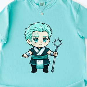 Peut inclure: T-shirt bleu clair avec un personnage de dessin animé aux cheveux turquoise et aux yeux bleus. Il porte une tenue bleu foncé et blanche et tient un bâton argenté. Le t-shirt a une étiquette Comfort Colors.