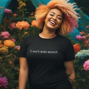 Peut inclure: T-shirt noir avec le texte "I can't mute myself." Une personne aux cheveux bouclés orange et jaune sourit devant un jardin fleuri coloré. Les fleurs comprennent des fleurs roses, oranges et bleues.