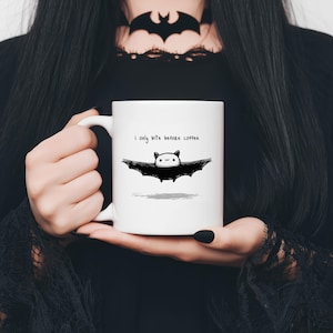 Puede incluir: Taza de cerámica blanca con una ilustración de murciélago en blanco y negro y el texto "i only bite before coffee". El murciélago tiene una cara linda. La taza es sostenida por una persona con una parte superior negra.