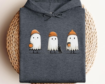 May include: Dark grey hoodie with a playful design of three cartoon ghosts in orange beanies. One ghost holds a pumpkin, another a black cat, and the third a coffee cup. The image is printed on the folded hoodie.