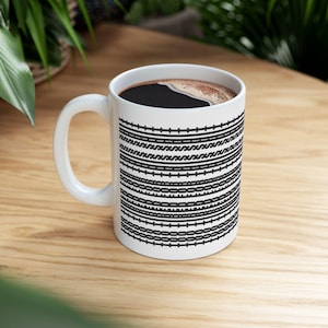 May include: White ceramic coffee mug with a black geometric pattern of horizontal lines and shapes. The mug is filled with dark coffee and has a white handle. The mug is on a wooden surface.