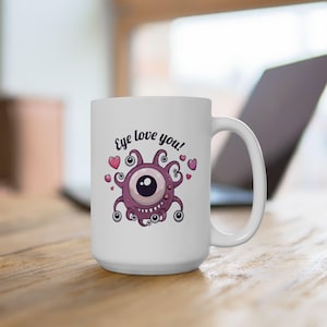 May include: White ceramic mug featuring a cartoon monster with a large eye, tentacles, and hearts. The mug has the text "Eye love you!" in a whimsical font. The mug is on a wooden surface.
