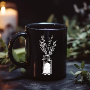 May include: Black ceramic mug with a white illustration of herbs in a jar. The mug has a curved handle and a smooth surface. The design features sprigs of lavender and rosemary. The mug is set against a dark background with candles and greenery.
