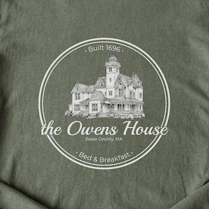 May include: Olive green t-shirt with a white circular design. The design includes a detailed illustration of a Victorian house, the text "the Owens House", "Essex County, MA", and "Bed & Breakfast". Above the house is the text "Built 1696."