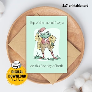 May include: A 5x7 printable greeting card with a light green background featuring an illustration of a frog wearing a plaid jacket, vest, and hat. The text reads Top of the mornin' to ya on this fine day of birth. The card is a digital download.