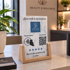 Peut inclure: Un porte-affiche en acrylique transparent avec une base en bois clair présentant une carte à motif marbre blanc. La carte affiche le logo SFrame, une icône NFC, un code QR, cinq étoiles bleues et le texte Lascia anche tu una recensione et dove le idee prendono vita.
