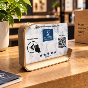 Pode incluir: Um suporte de madeira clara curvada segurando uma placa retangular com padrão de mármore branco. A placa apresenta o texto Lascia anche tu una recensione, SFRAME Graphic Designers, Tap your phone, Scan QR-code, um ícone NFC e cinco estrelas azuis.