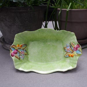 May include: A green ceramic serving dish with a scalloped edge and floral designs. The dish features painted flowers and berries in shades of pink, red, yellow, and green.