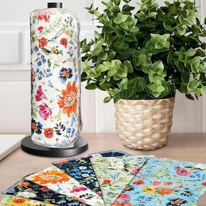 Puede incluir: Un rollo de papel de cocina con estampado floral sobre un soporte negro, con una variedad de paños con estampado floral a juego abanicados debajo. Una planta en maceta en una cesta tejida está a la derecha.