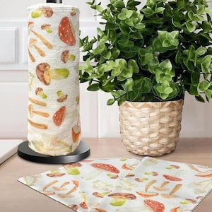 May include: A roll of paper towels with a mushroom design stands on a black base. The towels feature illustrations of various mushrooms in shades of red, brown, and green. A small potted plant in a woven basket and a matching cloth are also visible.