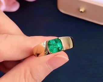 May include: A gold ring featuring a large, square-cut emerald gemstone. The ring has a simple, modern design with a polished finish. The emerald is a vibrant green color, set in a gold band. The ring is held up against a dark blue background.