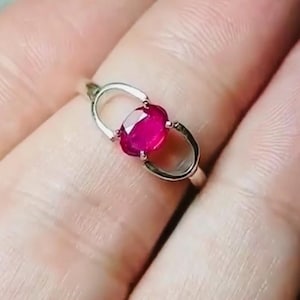 Peut inclure: Une bague en argent ornée d'un rubis ovale d'un rouge vif. Le rubis est serti dans un design unique à cadre ouvert, mettant en valeur sa couleur rouge intense. L'anneau est fin, soulignant la proéminence du bijou.
