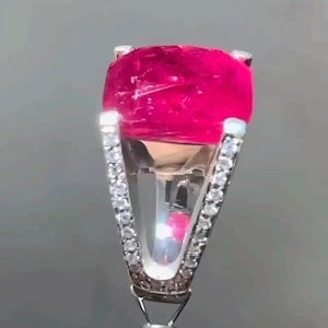 May include: A close-up of a ring featuring a large, vibrant pink gemstone. The ring's band is silver and adorned with small, sparkling clear stones. The gemstone is a vivid, saturated pink color, set in a classic prong setting.