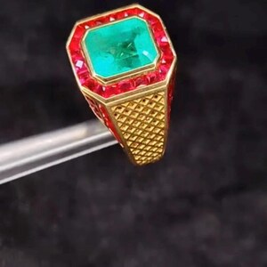 May include: A gold ring featuring a large, rectangular emerald center stone surrounded by a border of small red gemstones. The ring's band has a textured, grid-like pattern.