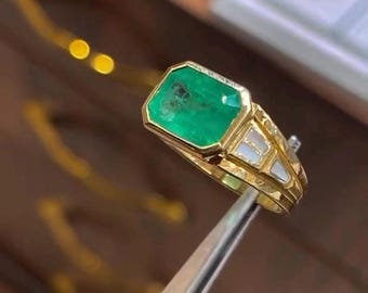 May include: A gold ring with a large, rectangular emerald gemstone. The ring has a detailed design with white accents and is held by a pair of silver tweezers. The ring is a piece of jewelry.