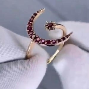May include: A gold-colored ring featuring a crescent moon and star design. The moon is adorned with small, round, red gemstones. The star is also embellished with red gemstones. The ring is set against a soft, neutral background.