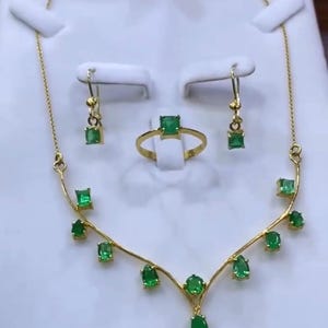 May include: A gold jewelry set featuring a necklace, earrings, and a ring, all adorned with vibrant green emeralds. The necklace and earrings have a delicate design, while the ring showcases a square-cut emerald.
