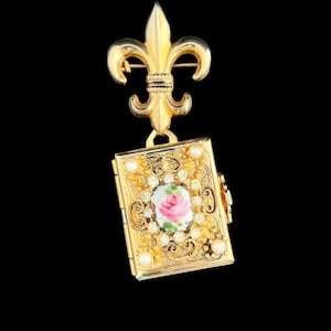 May include: A gold-toned brooch featuring a fleur-de-lis design at the top, connected to a small, rectangular locket. The locket is adorned with a central pink rose design, surrounded by clear crystals and small pearls. The brooch has an ornate, vintage aesthetic.