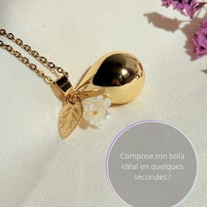 Puede incluir: Un collar con colgante dorado con un dije en forma de pera, una pequeña flor blanca y una hoja. El collar tiene una cadena fina. La imagen también incluye texto en un círculo.