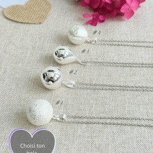 Può includere: Quattro collane bola color argento con diversi design a sfera sono esposte. Le collane sono su un tessuto beige chiaro, con fiori secchi e decorazioni a forma di cuore sullo sfondo. Il testo "Choisi ton bola" è visibile.