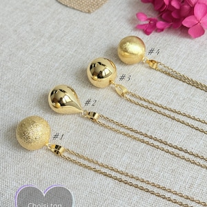 Può includere: Quattro collane bola color oro con diversi design di ciondoli. Ogni collana ha una catena delicata e un'etichetta numerata. I ciondoli includono una sfera strutturata, una forma a goccia, una sfera con ritagli e una sfera liscia. L'immagine include anche il testo "Choisi ton bola".