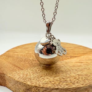 Peut inclure: Un collier argenté avec un pendentif sphérique, un cœur et un charm en forme d'ours. La chaîne est argentée. Le pendentif reflète l'environnement. Le collier est présenté sur une surface en bois.