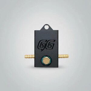 May include: A black rectangular device with the letters "GG" engraved on the front. It has two brass tubes extending from the sides and a circular gold-colored element in the center. A small loop is at the top.