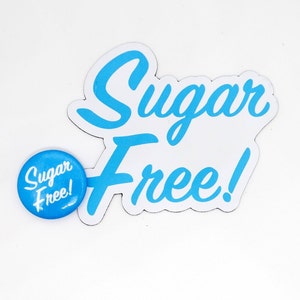 May include: A blue and white sticker with the text "Sugar Free!" in a cursive font. A smaller blue button with the text "Sugar Free!" in white is also shown.