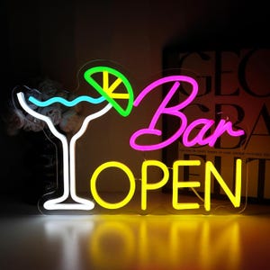 Op de afbeelding: Een neonbord met een cocktailglas en de woorden "Bar OPEN". De cocktail is wit omlijnd en gevuld met blauw, met een groene en gele limoenpartje. Het woord "Bar" is roze en "OPEN" is geel.