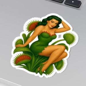 Puede incluir: Una pegatina con una ilustración estilo pin-up de una mujer con cabello verde y un vestido verde, sentada entre trampas para moscas Venus. El vestido tiene un ribete rojo y las trampas para moscas son verdes con interiores rojos.