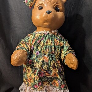 May include: A brown teddy bear wearing a green dress with a floral print and a matching bow. The dress has a white lace trim.