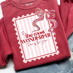 May include: A maroon sweatshirt with the text "It's the most WONDERFUL Time of the year" in a stamp-like design. The sweatshirt is folded with a pair of blue jeans on a white background.