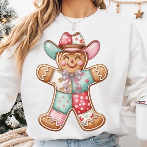 Könnte beinhalten: Weißes Sweatshirt mit einem Lebkuchenmann mit Cowboyhut. Der Lebkuchenmann hat ein Patchwork-Design mit rosa, blauen und roten Abschnitten. Eine Weihnachtstasse mit dem Weihnachtsmann wird in der Hand gehalten.